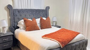 A bedroom with a tufted headboard, a bed with white and brown bedding, a brown throw blanket, a nightstand with a lamp, and a dark rug. - Modern Apartment Comfortable & Quiet (Santo Domingo Este)