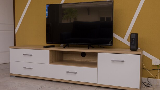 Een televisie op een houten en witte kast met lades, voor een gele muur met witte lijnen.