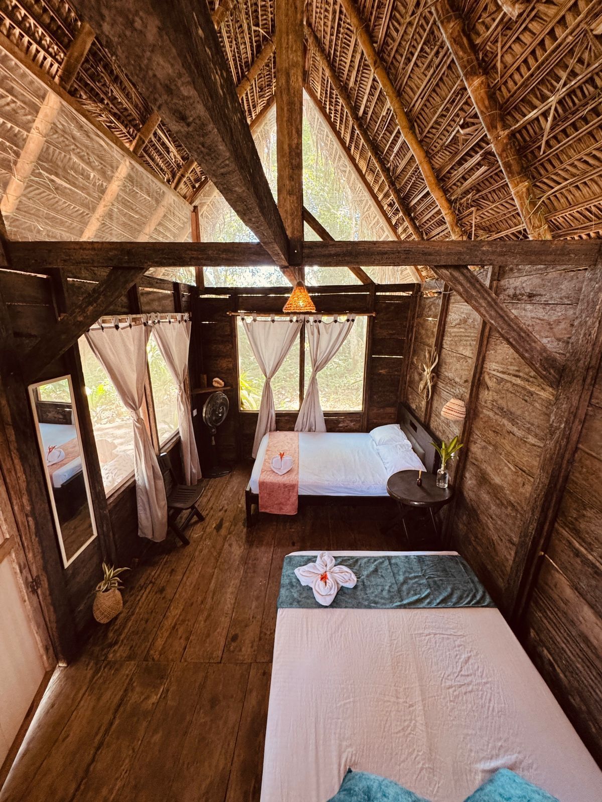 A rustic wooden cabin with two beds, a fan, and a window with curtains.