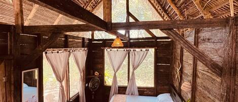 A wooden cabin with a thatched roof, two beds, a small table, and a fan.