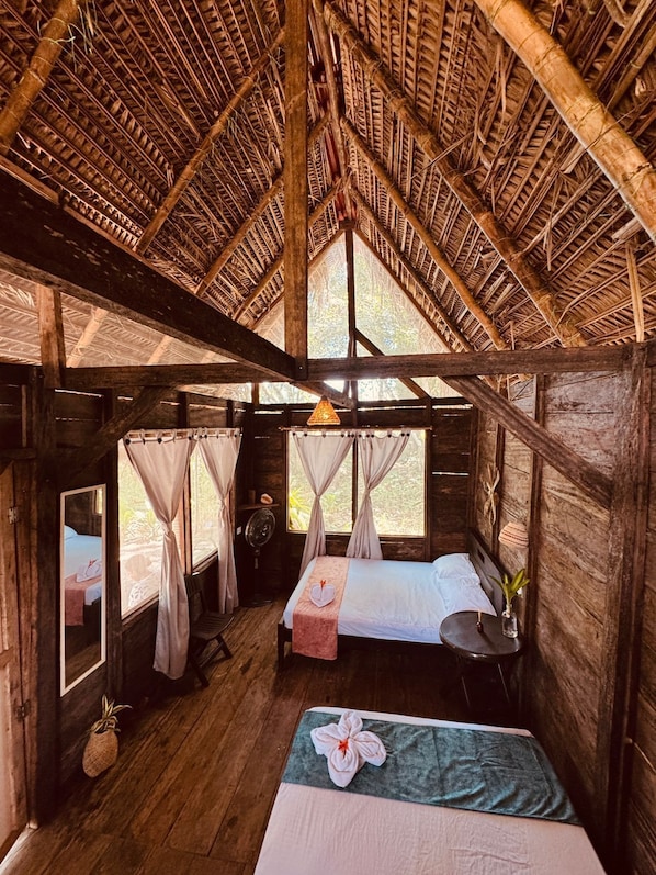 Eine Holzhütte mit einem Reetdach, zwei Betten, einem kleinen Tisch und einem Ventilator.