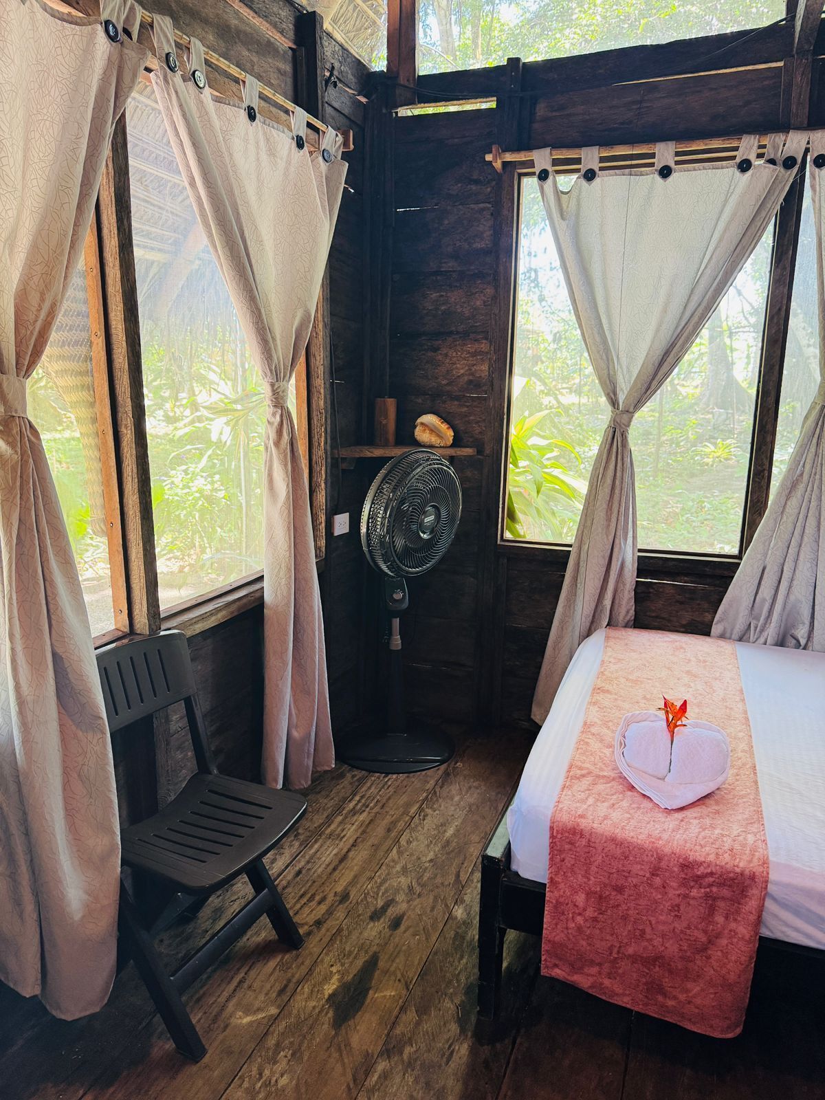 Quarto de madeira com cama, cadeira, ventoinha e cortinas.