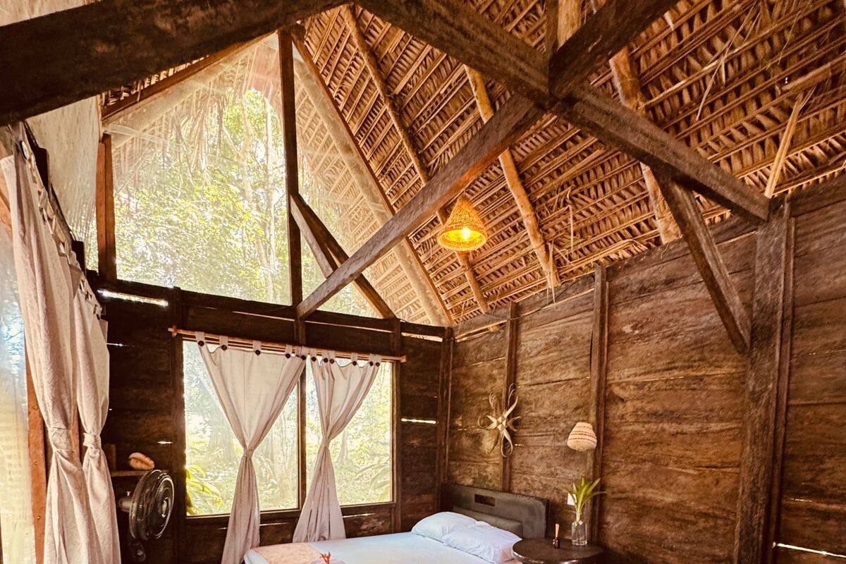 Una habitación rústica de madera con dos camas, un ventilador, una mesita y una ventana con cortinas.