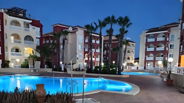 A swimming pool with a tiled deck, surrounded by palm trees and buildings with balconies. - Puerto Marina Apartment with Mountain Views, Wi-Fi, and Air Conditioning (Los Alcázares)