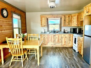 A kitchen with wooden cabinets, a dining table with chairs, a clock on the wall, and a window with a view. - Outdoorsman's Paradise with the Rio Grande National Forest as your backyard! (La jara)