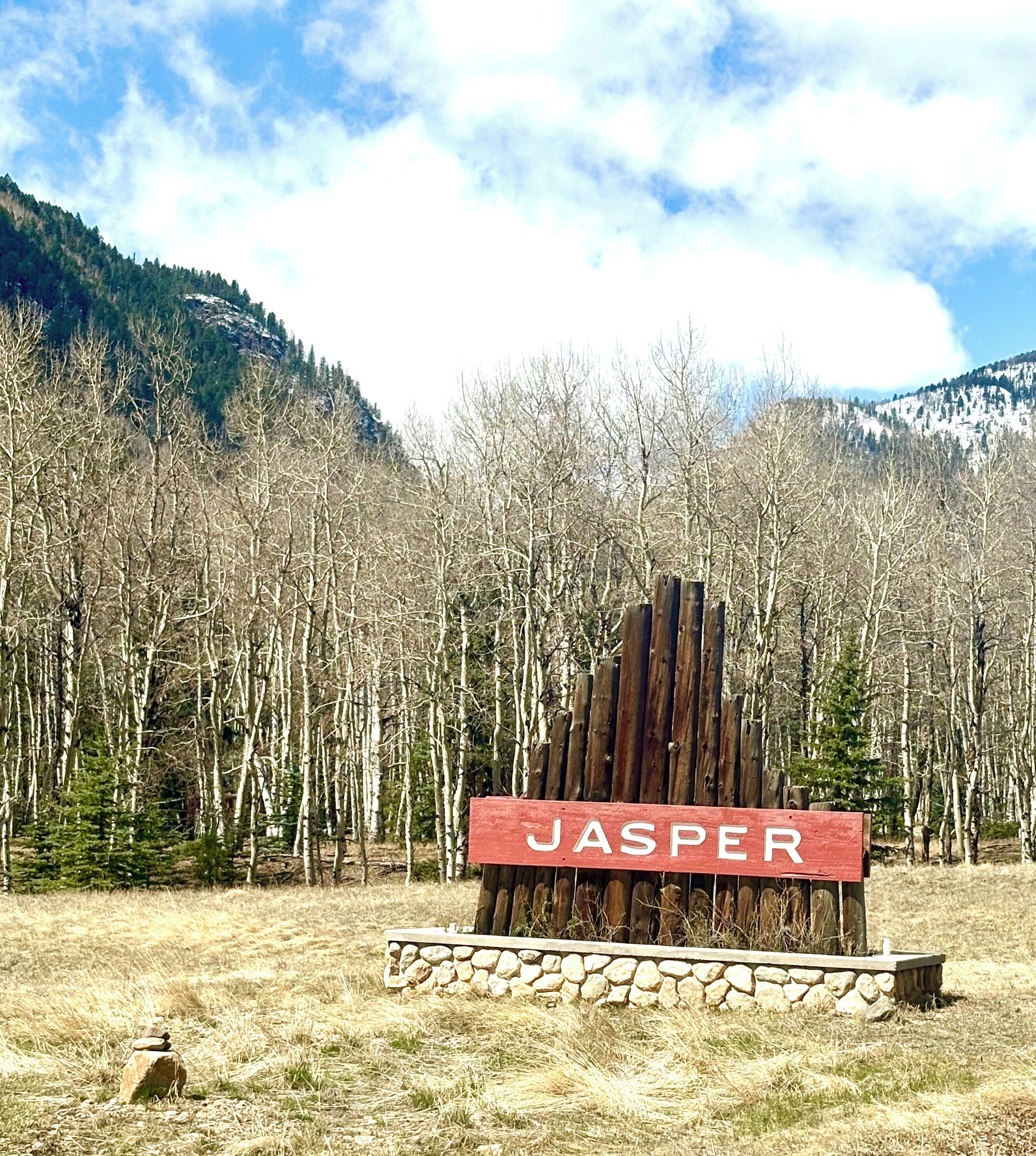 Un letrero de madera con la palabra 'JASPER' en letras blancas sobre un fondo rojo, frente a un paisaje de bosque y montañas.