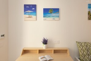 Une pièce avec un bureau en bois, un vase blanc orné de fleurs violettes et deux tableaux sur le thème de la plage accrochés au mur.