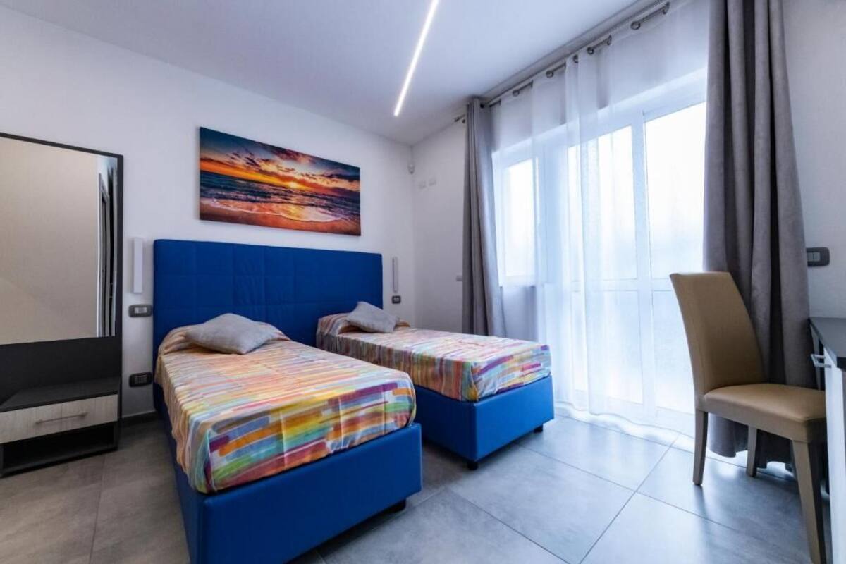 Ein Hotelzimmer mit zwei Betten, einem Sessel, einem Schreibtisch, einem Gemälde und einem Fenster mit Vorhängen.