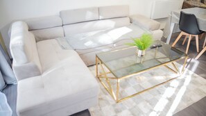 Un salon moderne avec un canapé d’angle, une table basse en verre aux pieds dorés, une petite plante en pot et un coin repas comprenant une table et des chaises.