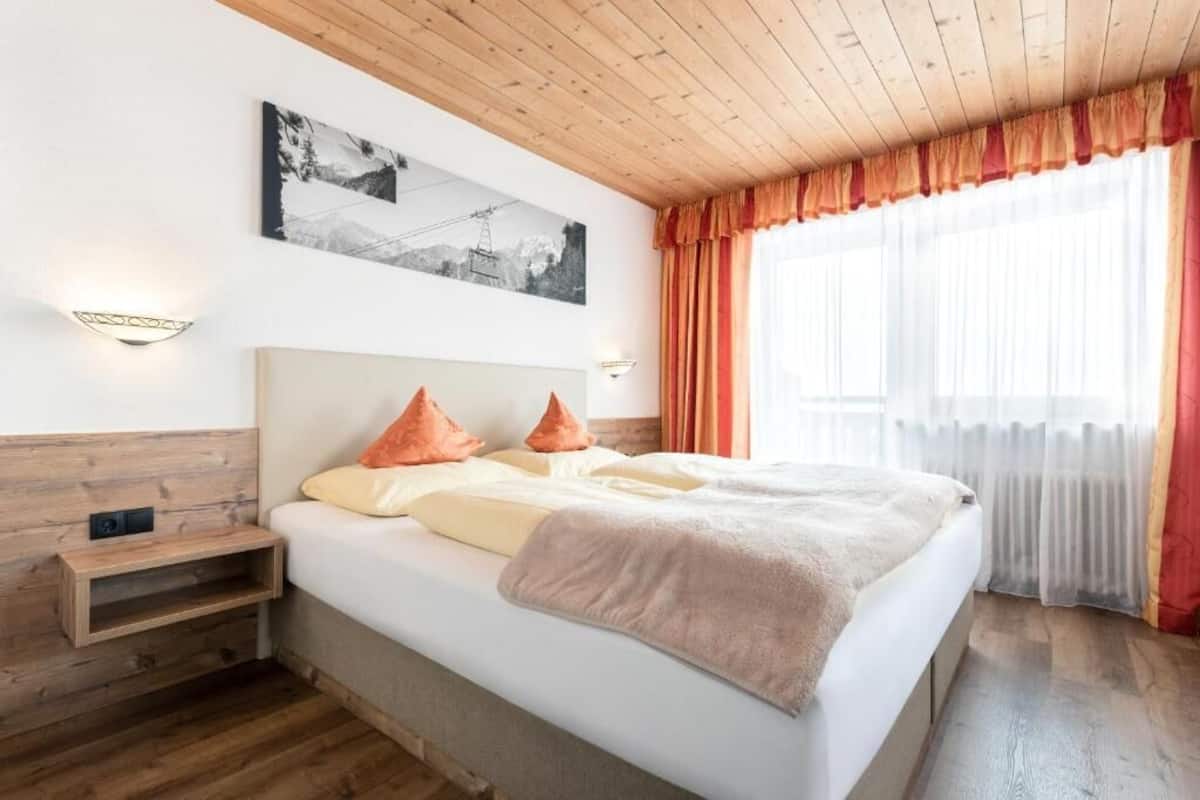 Un dormitorio con cabecera de madera, una cama con almohadas, una mesita de noche, una lámpara de pared, una ventana con cortinas e imagen en la pared.