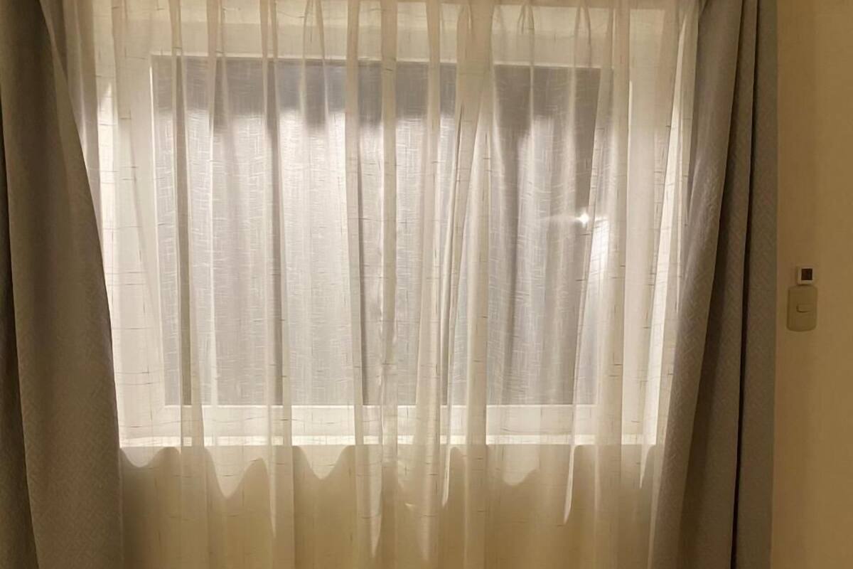 Una habitación con suelo de madera, una ventana con cortinas translúcidas y una barra para cortinas de madera.