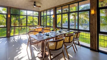 A spacious dining area with a large table and chairs, large windows with a view of greenery, and ceiling fans.