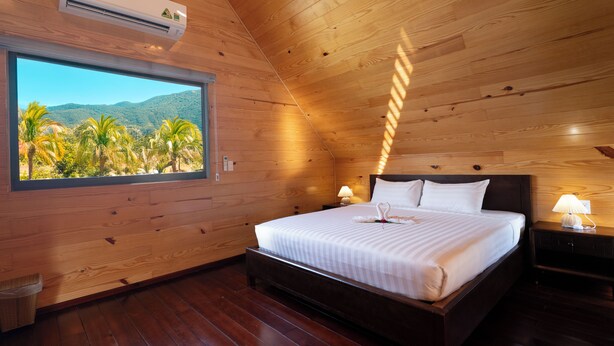 Una habitación con paneles de madera, una cama, una ventana con vista a palmeras y montañas, y un equipo de aire acondicionado.