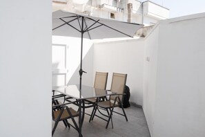 Une petite terrasse extérieure avec une table et des chaises en verre, un parasol noir et un mur blanc.