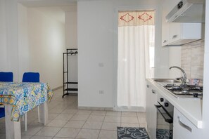 A compact kitchen with a gas stove, sink, and white cabinets. There is a dining table with a blue tablecloth and chairs. - Apartment 'Adriatic Breeze' with Private Terrace and Air Conditioning (Melendugno)