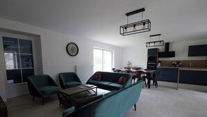 A modern living room with a sofa, chairs, a coffee table, and a dining area with a table and chairs. There is a kitchen area with cabinets and appliances.