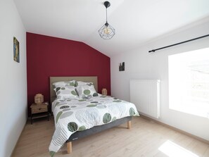 A bedroom with a bed, a red accent wall, a hanging light fixture, and a window.