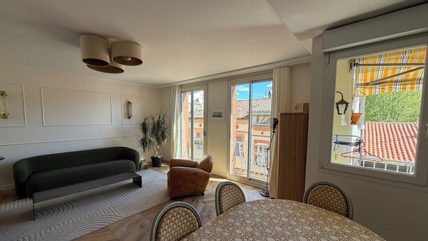 Ein Wohnzimmer mit einer Couch, einem Esstisch mit Stühlen, einer Zimmerpflanze und Blick auf einen Balkon mit Geländer.