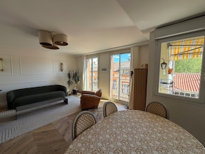 Een woonkamer met een bank, een eettafel met stoelen, een potplant en uitzicht op een balkon met een hekwerk.
