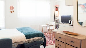 Habitación de hotel con cama, mesita de noche, escritorio, silla, armario, espejo y ventana con cortinas.