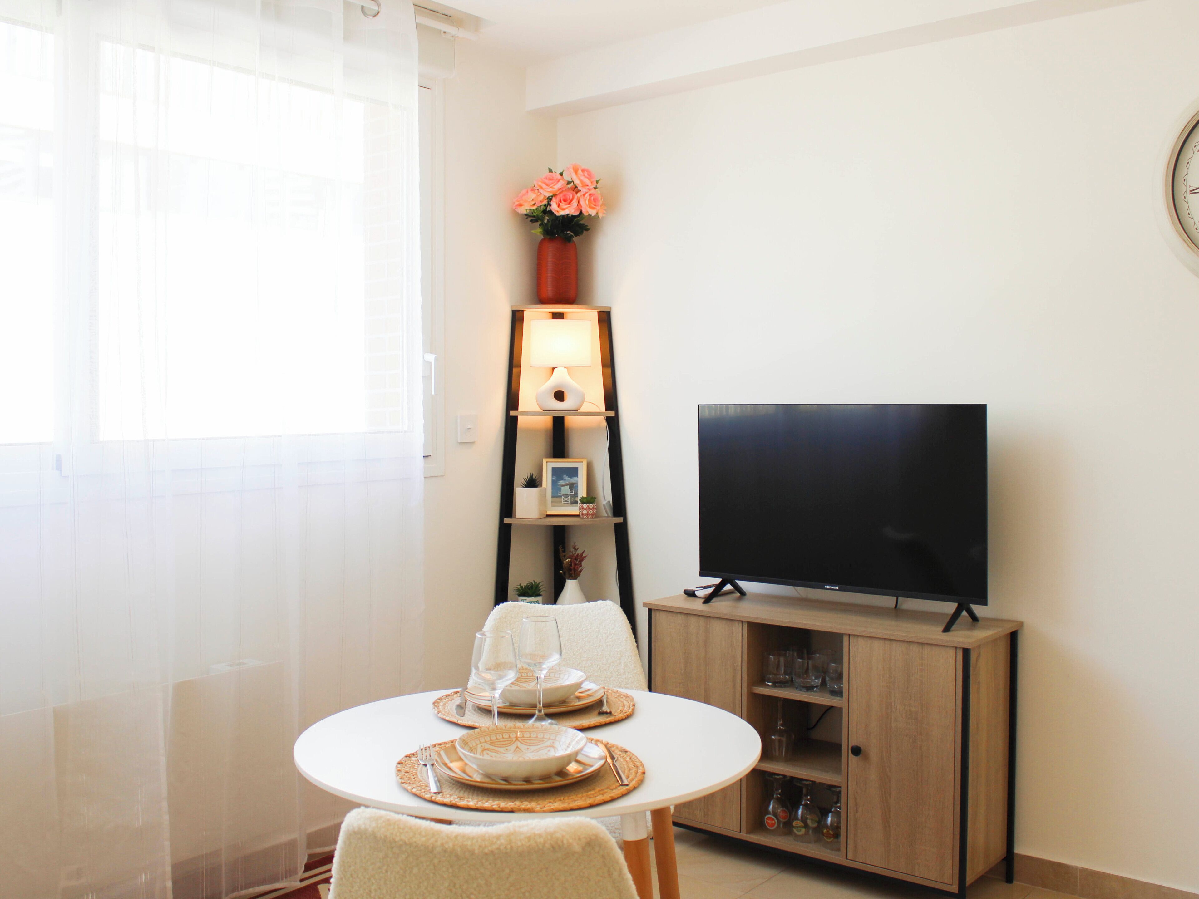Uma sala moderna com televisão de tela plana, uma mesa de jantar com jogos americanos, um vaso com flores e uma luminária em uma estante.