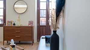 Ein Zimmer mit einem Holzkleiderschrank, einem runden Spiegel, einer Vase mit Trockenpflanzen und einem Fenster mit Blick auf Gebäude.