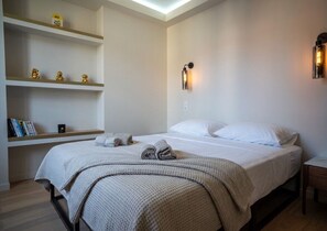 Un letto rifatto con un copriletto bianco e cuscini, un comodino con una lampada e mensole integrate con oggetti decorativi.