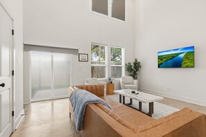 Un salón espacioso con un gran ventanal corredera, un sofá de cuero marrón, un sillón blanco, una mesita auxiliar y un televisor de pantalla plana montado en la pared.