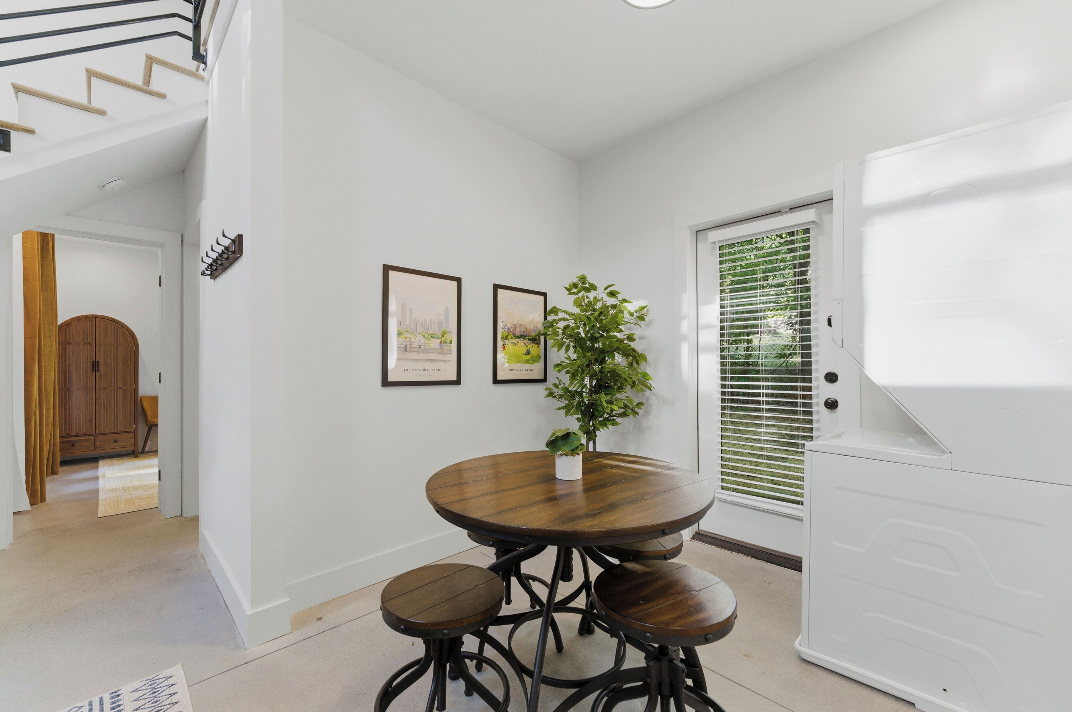 A dining area with a round table and stools, a potted plant, framed pictures on the wall, and a white appliance.