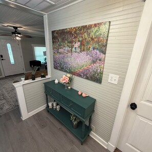 A room with a green console table, a large floral painting, and a ceiling fan. - The 1918 Magnolia Cottage (Milton)