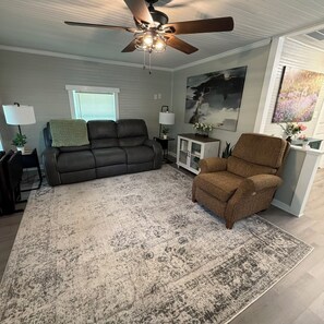 A living room with a ceiling fan, a black leather sofa, a brown armchair, a patterned rug, and a white cabinet with a vase of flowers. - The 1918 Magnolia Cottage (Milton)
