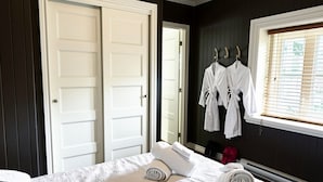 A hotel room with a bed, two white doors, a window with blinds, and a robe hanging on the wall.