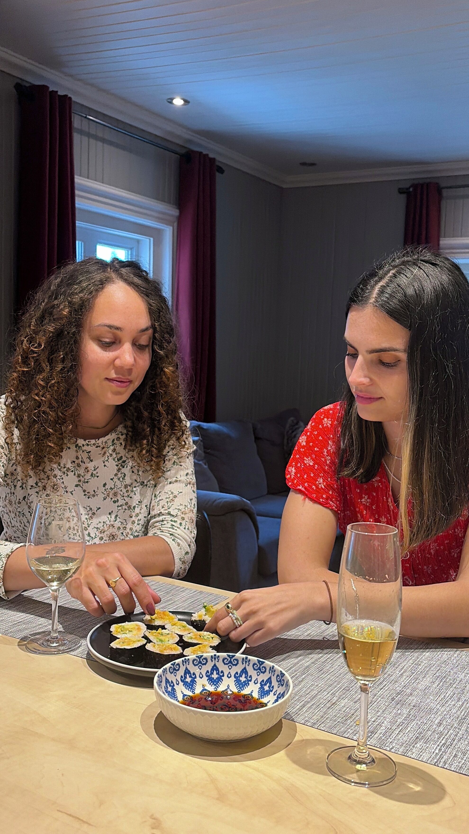 Duas pessoas sentadas à mesa com um prato de comida e copos de um líquido transparente, provavelmente vinho, num espaço com um sofá e cortinas ao fundo.