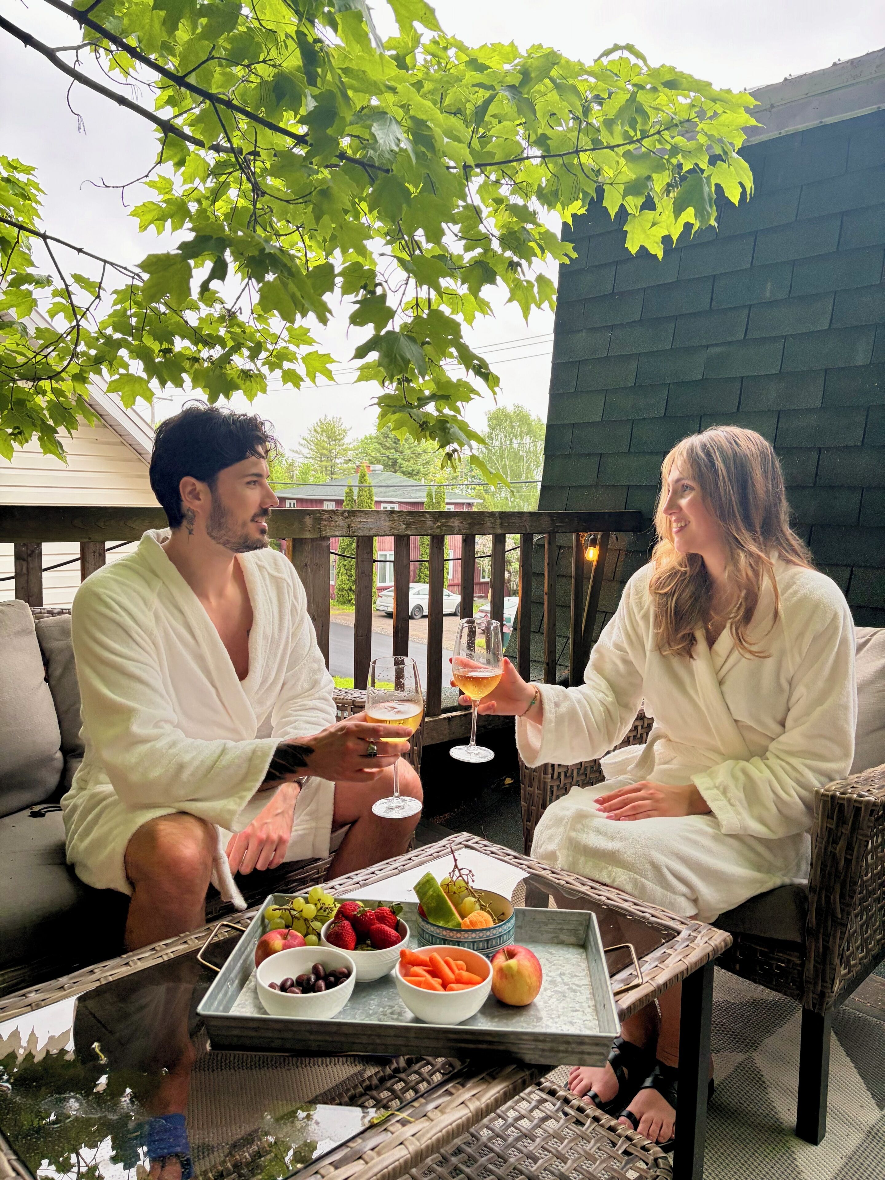 Two people in white robes sitting on a wicker chair outdoors with a tray of fruits and drinks in front of them.