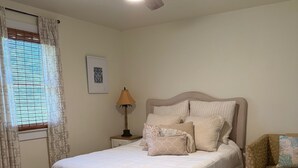 Une chambre à coucher comprenant un lit, une table de chevet, une lampe, un ventilateur de plafond et une fenêtre avec des rideaux.