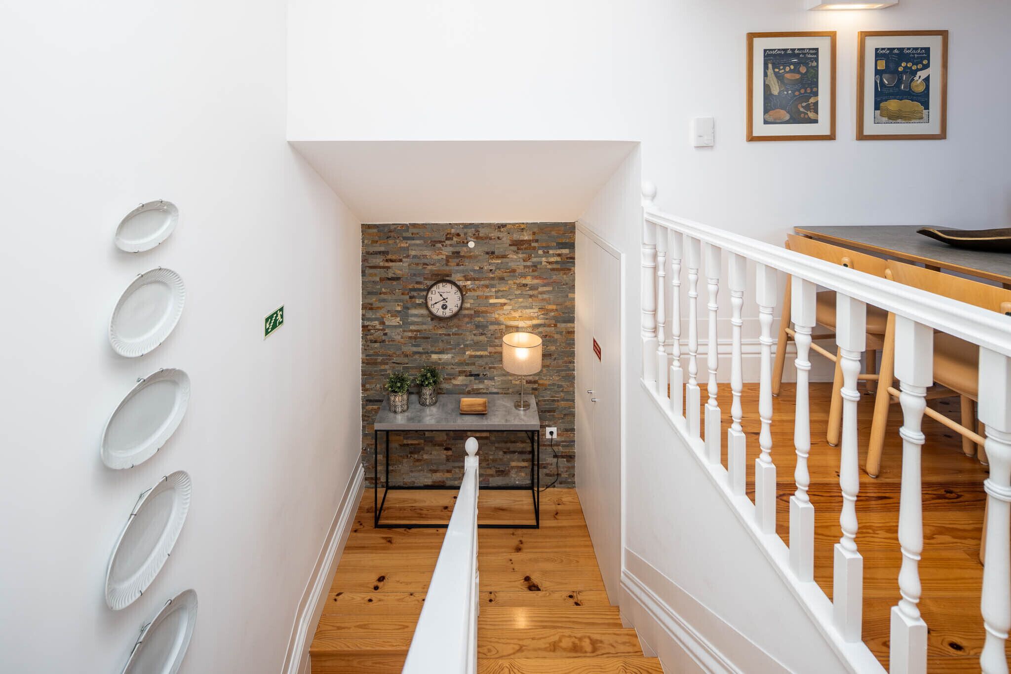 A staircase with a white railing, a wooden floor, a wall with a clock, a table with a lamp, and framed pictures on the wall.