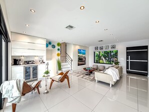 Una sala moderna con sofá, mesa de centro y sillas. Hay una zona de cocina con armarios y una mesa de comedor. La habitación tiene un ventanal con vistas a zonas verdes.