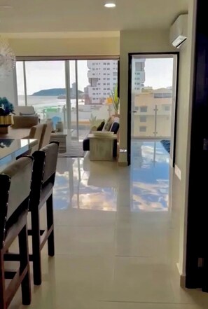 A modern interior with a dining area, a sofa, and a view of the city and sea through large windows. - Luxurious 2 bedroom condo with partial ocean view (Mazatlan)