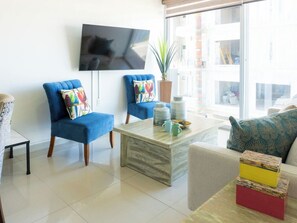 Een moderne woonkamer met twee blauwe fauteuils, een salontafel en een aan de muur bevestigde televisie.