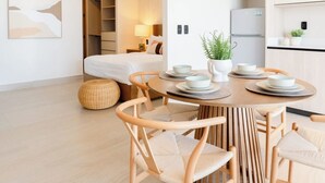 Ein modernes Hotelzimmer mit einem Bett, einer Essecke mit Tisch und Stühlen, einer Küchenzeile und einem Flur, der zum Badezimmer führt.