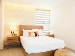 A neatly made bed with white bedding and multiple pillows, two bedside tables with lamps, a small potted plant, and a window with blinds. - Luxury apt on the Malecón with lagoon views (Mazatlan)