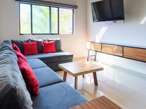 A modern living room with a sofa, a coffee table, a TV, and a window with blinds. - Family apartment with roof and great location (Mazatlan)