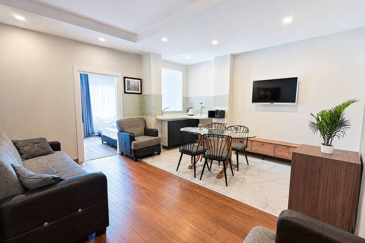 A modern living room with a sofa, a dining area with chairs, a kitchen with a sink, and a TV mounted on the wall.