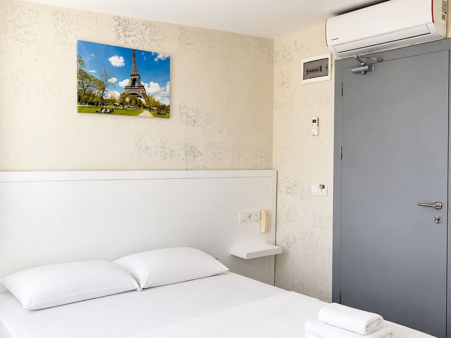 A hotel room with a bed, a wall-mounted air conditioner, a door, and a picture of the Eiffel Tower.