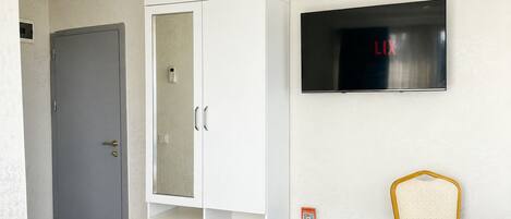 A room with a grey door, a white wardrobe, a small table with a chair, and a TV mounted on the wall.