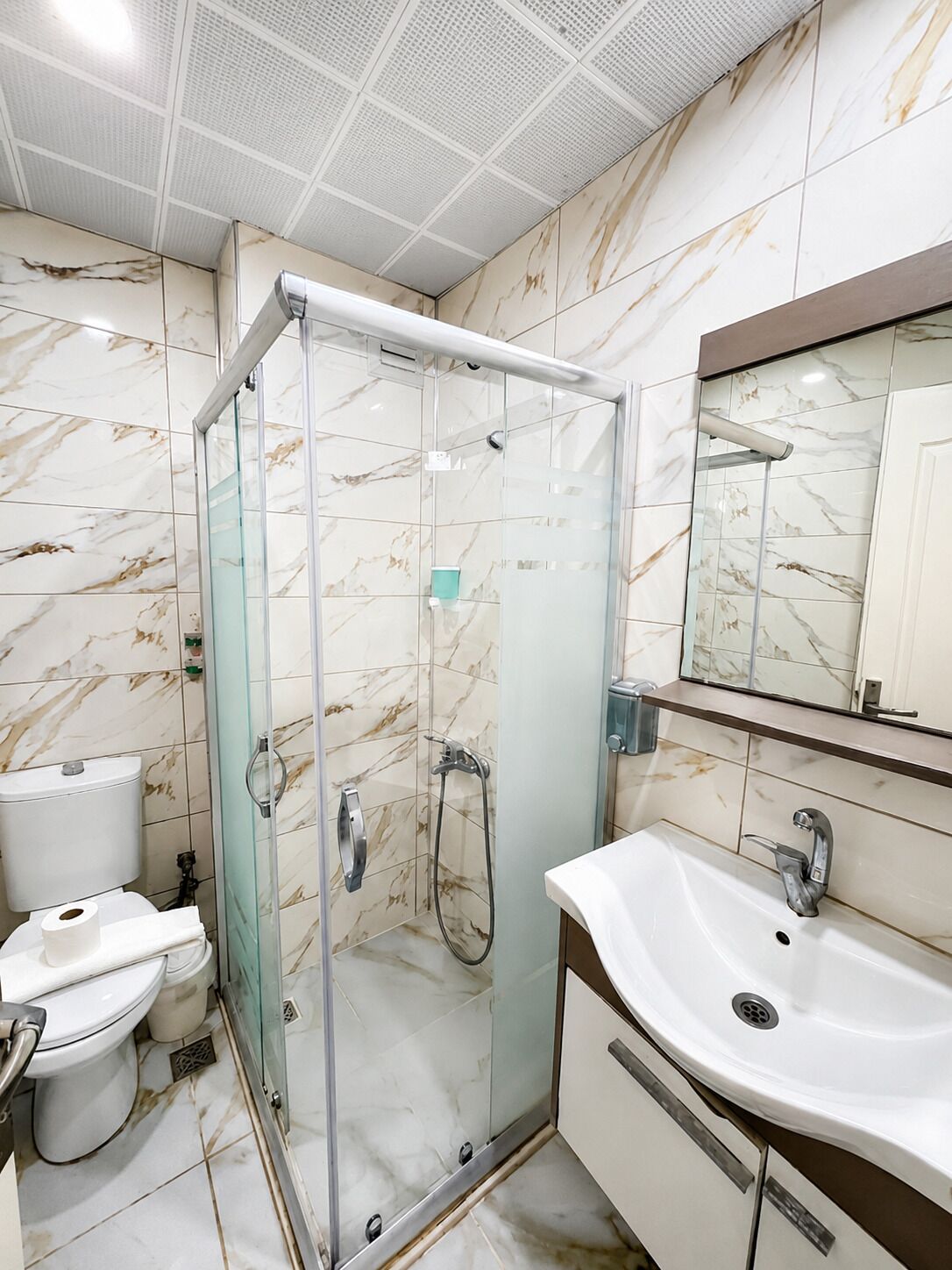 Un baño moderno con ducha de vidrio, un lavabo blanco y pared con azulejos de mármol.