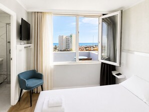 A hotel room with a bed, a TV, a chair, a window with a view of buildings and a beach. - Luna Hotel (Güzeloba)
