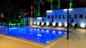 Una piscina con sillones de descanso y sombrillas, un cartel que dice 'HOTEL LUNA', y un entorno nocturno con árboles iluminados.