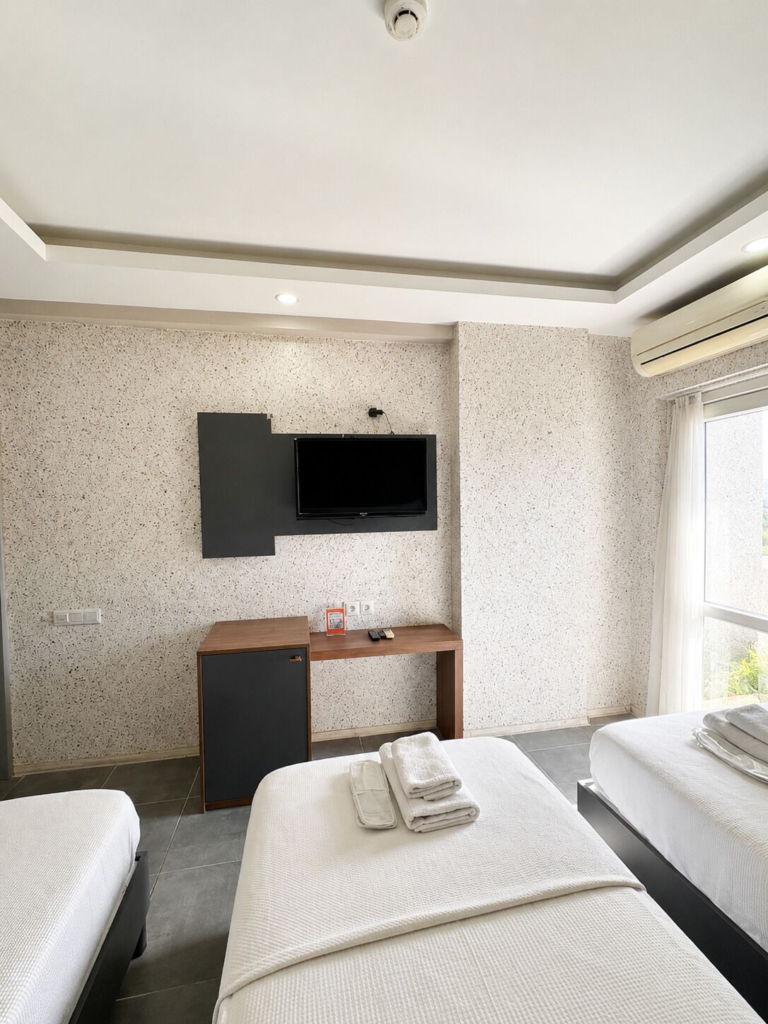 A hotel room with two beds, a TV mounted on the wall, a desk with a chair, and an air conditioning unit.