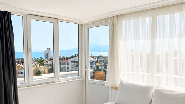 Habitación de hotel con cama, ventana que ofrece vistas a edificios y montañas, y una ventana con cortinas translúcidas.
